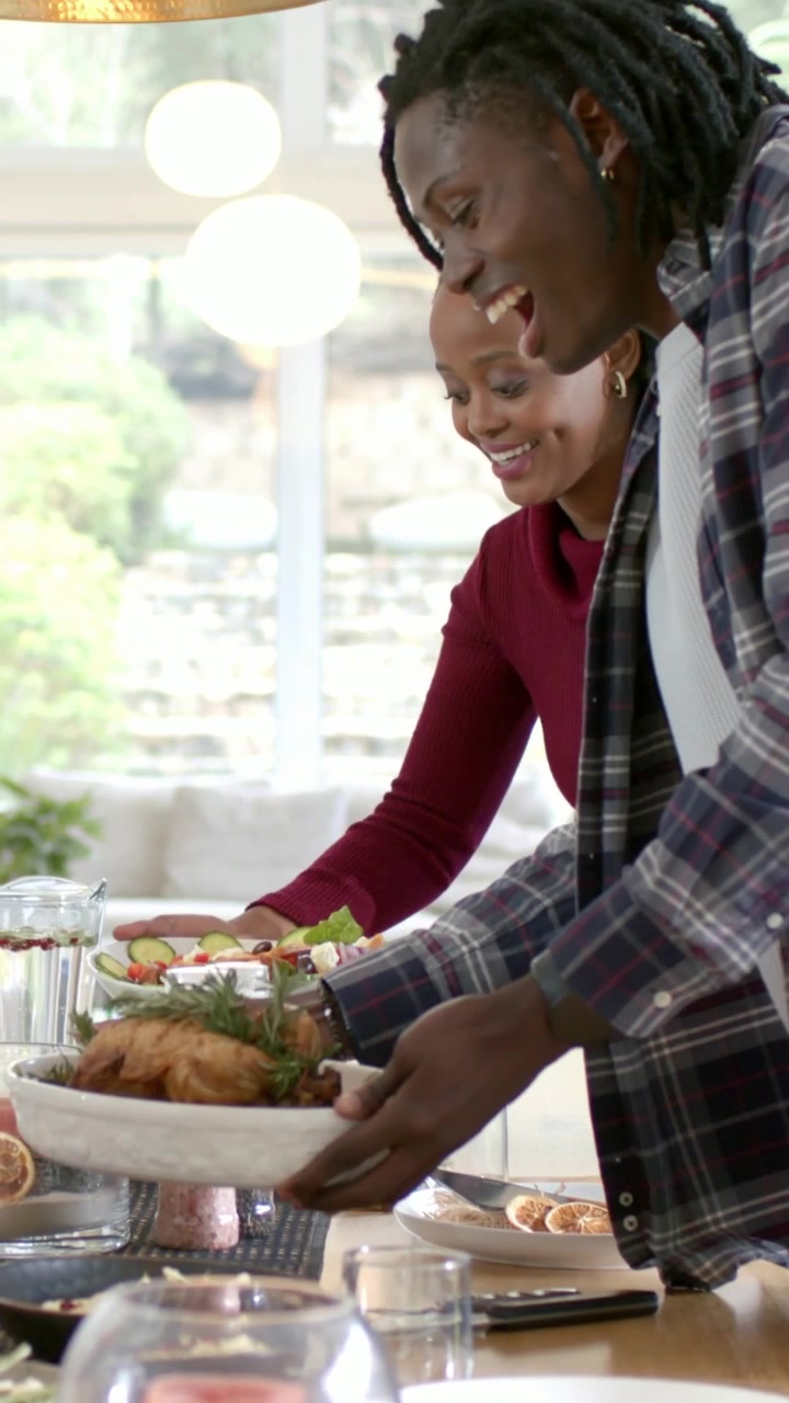 Vertical video: Entering maroon plaid couple placing roast and salad on dining table preparing meal