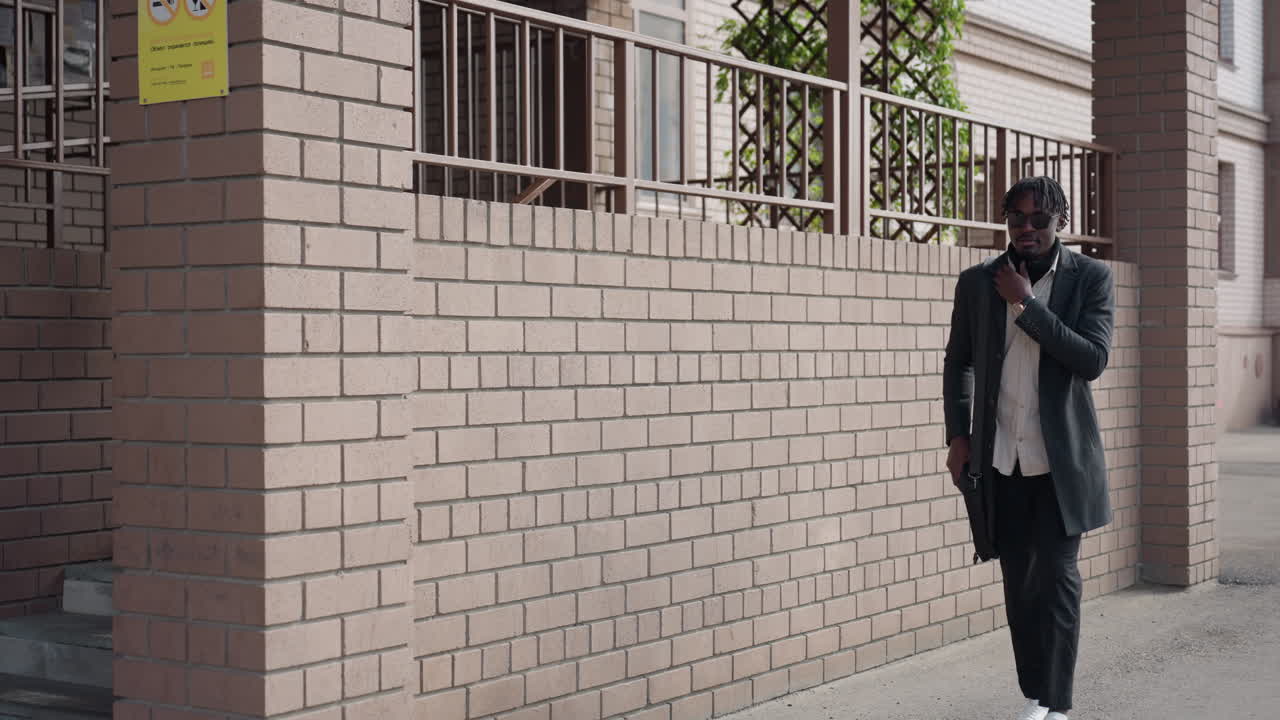 Professionals pass near brick entrance woman steps from corner as man approaches walkway, Sunglasses on, coats moving, Quiet city moment before work, sunlight hits wall while vines frame railing