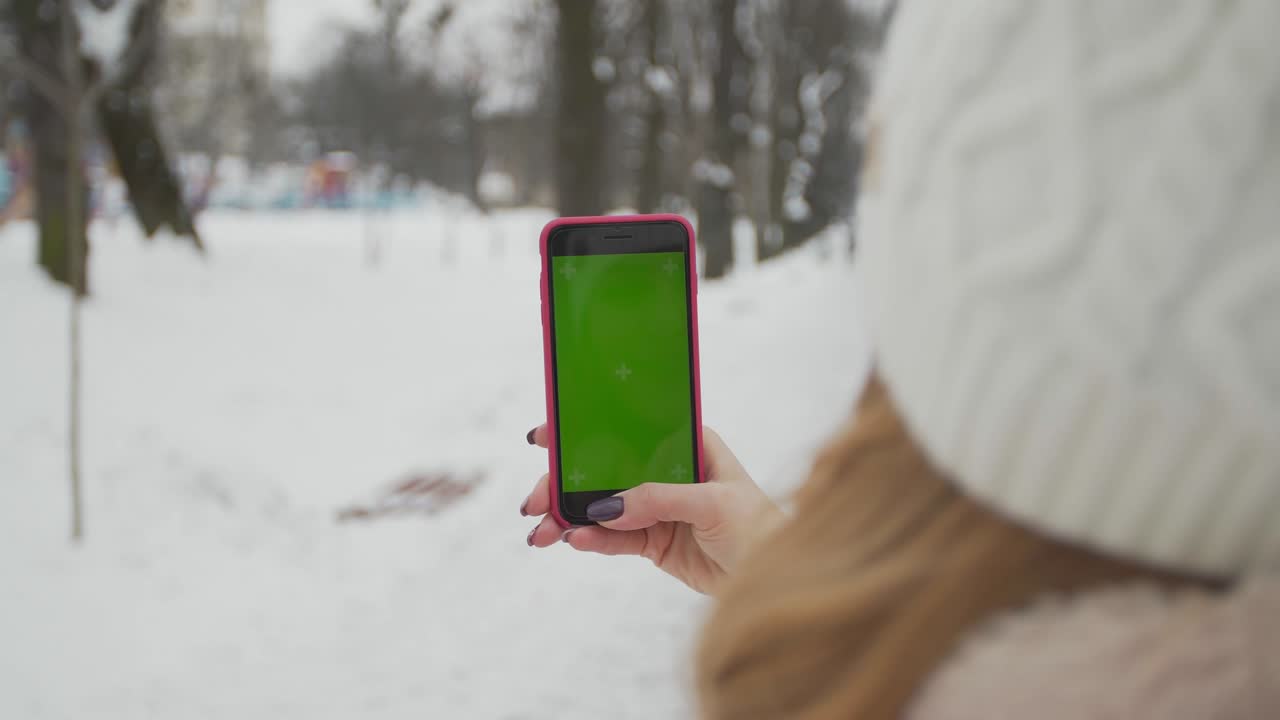 una mujer joven escribe y desliza en su teléfono inteligente en el parque de invierno. teléfono celular con pantalla de teclas cromadas - pantalla verde. cerrar. seguimiento de movimiento.