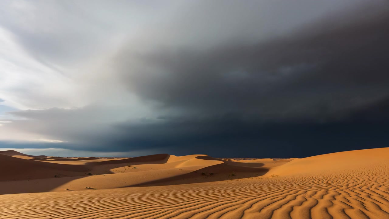 Ampio paesaggio arido con dune di sabbia ondulate in primo piano e un orizzonte basso e lontano sotto un cielo pieno di pesanti nuvole temporalesche. La luce naturale soffusa crea