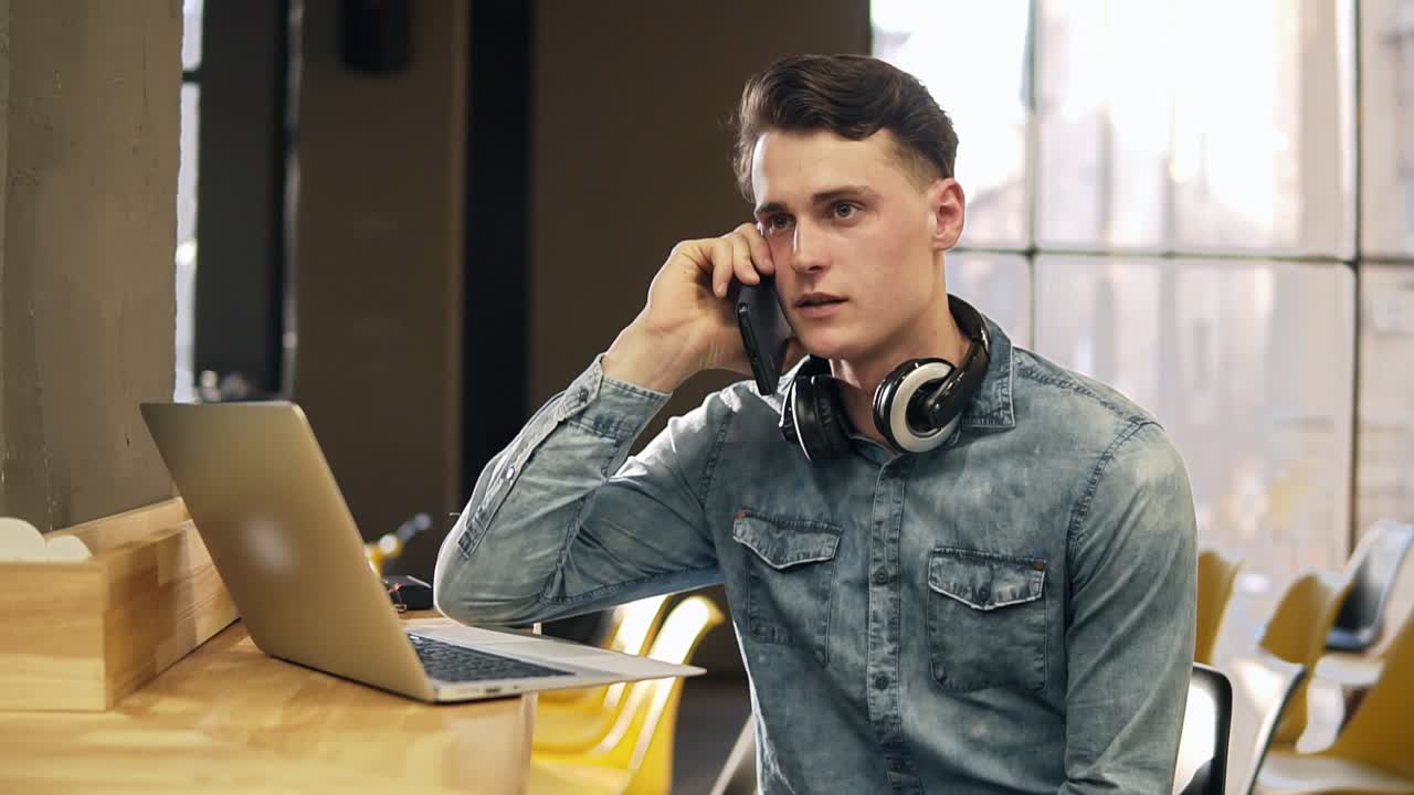 Attractive male in his 20's in denim shirt with wireless headphones around his neck and laptop beside him, talking with someone on the phone. Open co-working space. Indoors.