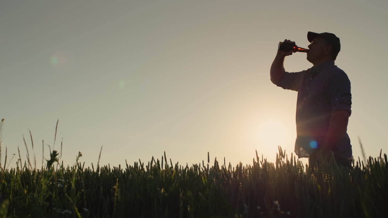 Sagoma di un uomo che beve birra sullo sfondo di un campo di grano dove il sole tramonta