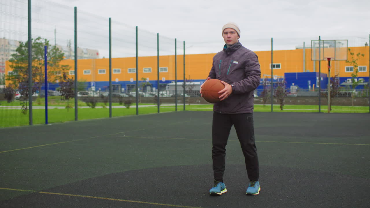 Homem branco praticando basquete em quadra, campo de esportes urbano cercado sob céu nublado, usando jaqueta e tênis, driblando, levantando para arremesso com salto, acompanhamento completo e foco constante, cidade