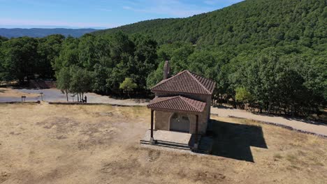 vuelo orbital sobre una ermita en un prado con un camino con una persona y su perro en un día de verano en un bosque frondoso con un valle con montañas en el fondo en ávila, españa