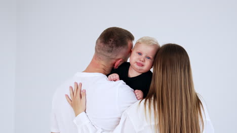 young caucasian parents stand their back to camera holding a baby. little kid kissed by mom and dad at white backdrop.