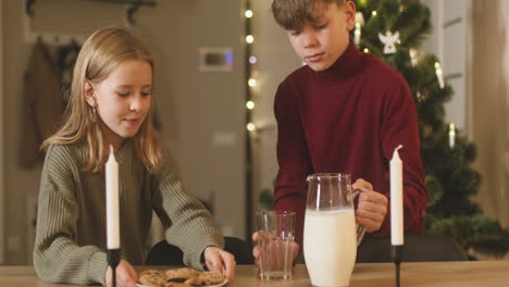 Un-Niño-Rubio-Y-Su-Hermana-Rubia-Colocan-Un-Vaso-Vacío,-Una-Jarra-De-Leche-Y-Un-Plato-Lleno-De-Galletas-En-Una-Mesa-Vacía-Con-Dos-Velas,-Luego-Salen-De-La-Habitación-1