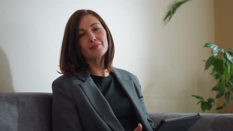 confident businesswoman in formal suit sitting on couch using digital tablet in modern office lounge surrounded by green indoor plants while reading or browsing calmly in natural daylight