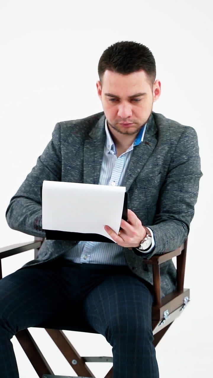 Handsome businessman reading documents. Young man in grey suit sitting on a wooden chair and looking the paperworks on white background. Vertical video