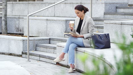 Mujer-De-Negocios-Sentada-Afuera-Durante-El-Almuerzo-Leyendo-Un-Libro-Y-Bebiendo-Café-Para-Llevar