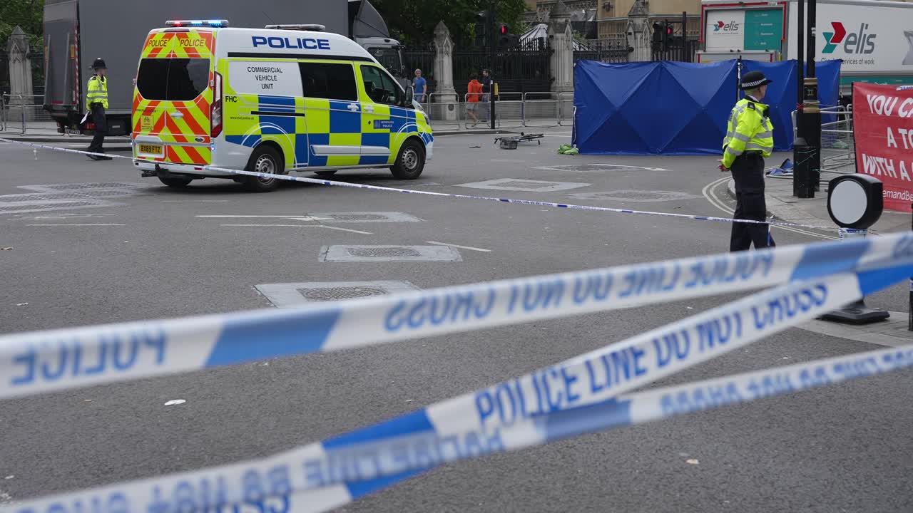A police officer stands behind blue and white cordon tape, a van is parked and blue tent surrounds a fallen bicycle at the scene vehicle collision in front of the Houses of Parliament.