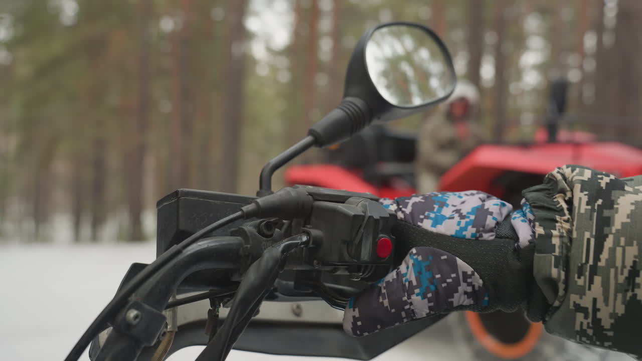 Primo piano del manubrio di un ATV con mano guantata, quad rosso sfocato in una foresta di pini innevata, blocco acceleratore e specchietto a fuoco, impugnatura testurizzata, atmosfera fredda e toni smorzati che trasmettono preparazione.