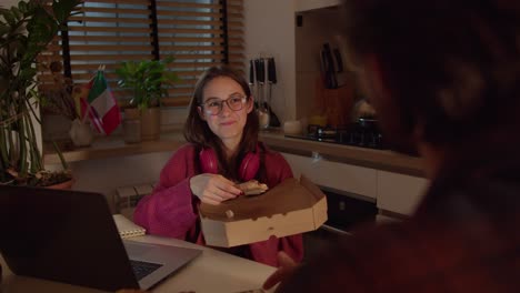 Confident-young-brunette-girl-with-glasses-in-red-wireless-headphones-and-a-sweater-holds-a-box-along-with-pizza-in-her-hands-and-makes-a-snack-during-her-foreign-language-lesson-with-a-teacher-in-the-kitchen-in-a-modern-apartment