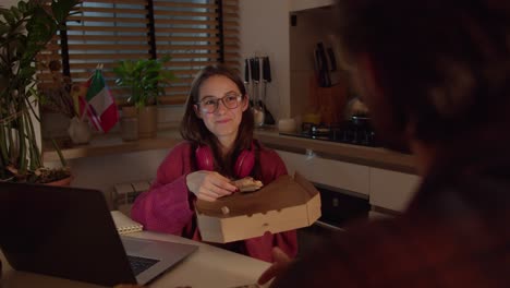 joven morena segura con gafas en auriculares rojos inalámbricos y un suéter sostiene una caja junto con pizza en sus manos y hace un bocadillo durante su lección de lengua extranjera con un maestro en la cocina en un apartamento moderno