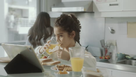 little girl using tablet on breakfast as mom cooking and talking on phone