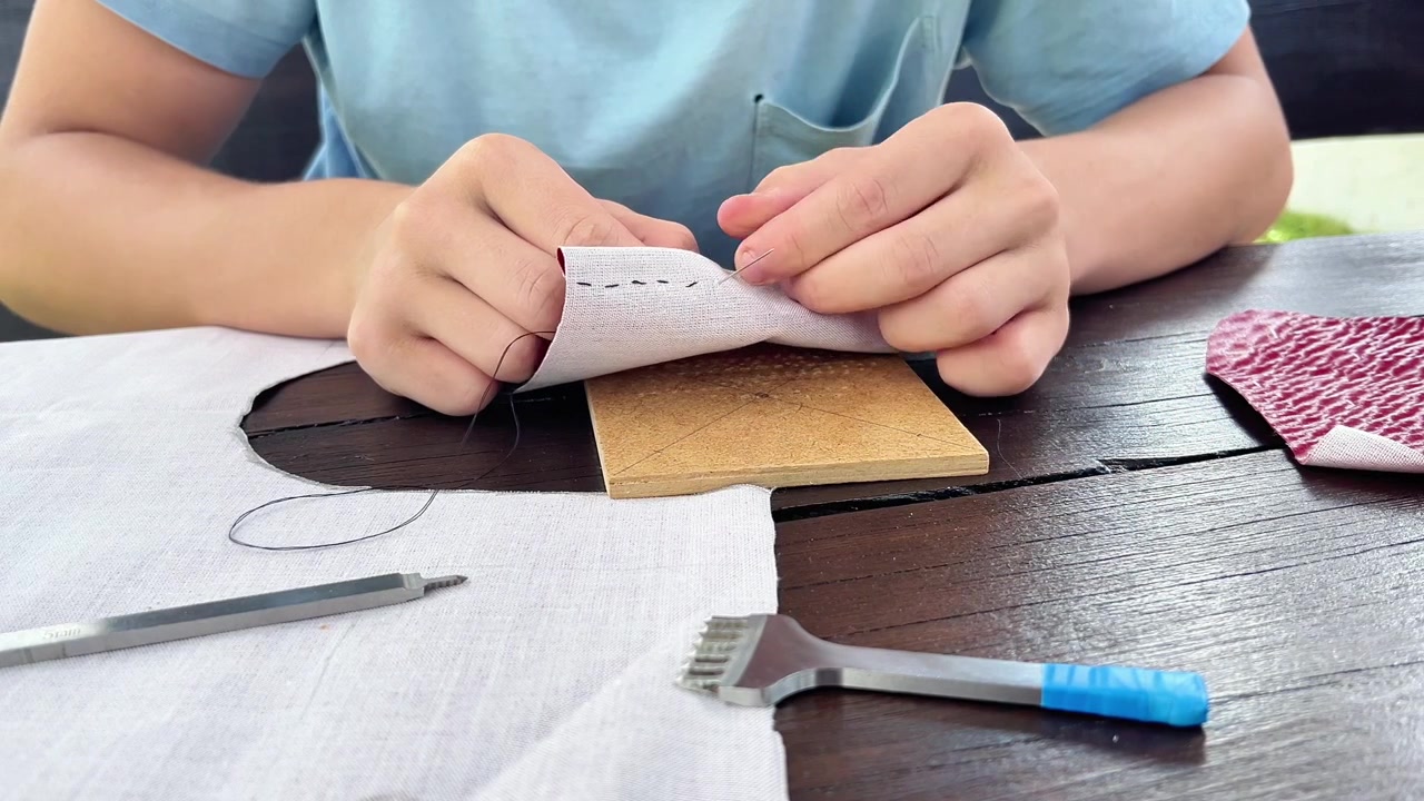 Close-up view of hands using a needle and thread to hand-sew leather and fabric at a workbench. Ideal for DIY tutorials, crafting tools, artisan marketing, and hobby content.
