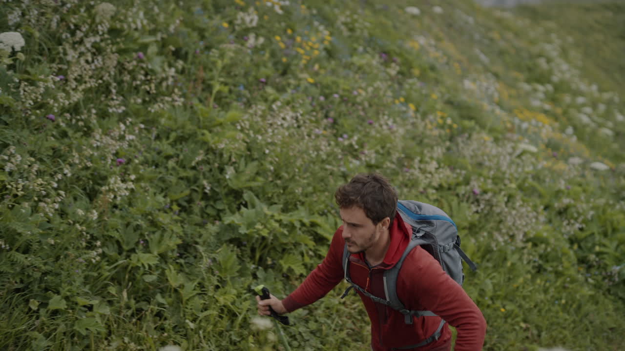 un joven excursionista con chaqueta roja y una mochila gris camina por un sendero hacia la cima de la montaña črna prst, donde hay una cabaña de montaña con bancos afuera para sentarse