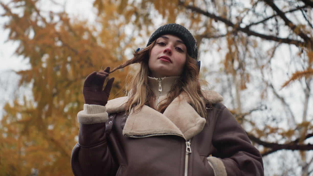 Beautiful lady with one hand in pocket and other hand rolling hair, wearing black knit cap and brown shearling jacket, enjoying autumn outdoors