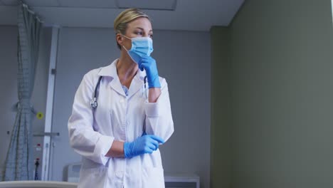 médica caucásica pensativa con máscara facial y guantes quirúrgicos de pie en el hospital