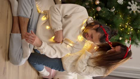 Vertical-video,-a-brunette-girl-in-a-White-sweater-decorates-her-brunette-boyfriend-in-a-red-New-Year's-hoop-with-the-help-of-a-yellow-glowing-garland-near-a-green-New-Year's-tree-in-a-cozy-room