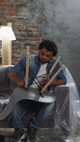 black man paints wall with roller in living room. african-american guy assembles chair legs sitting on sofa. renovation of apartment by roommates