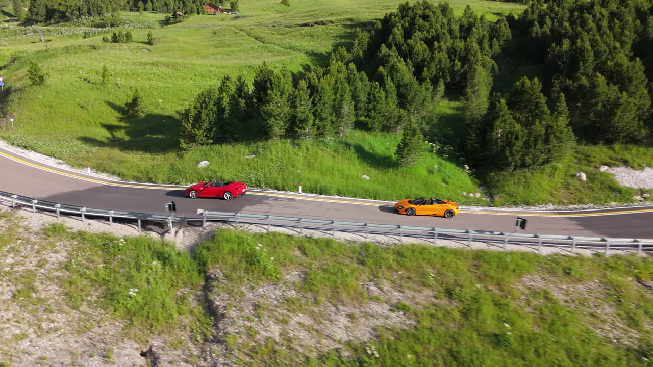 Dron śledzi dwa supersamochody na alpejskiej serpentynie, w otoczeniu lasów i szczytów