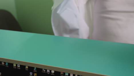 a close view of a man reaching for his lab coat, draped over a green counter, to wear it, with a green frame door behind him