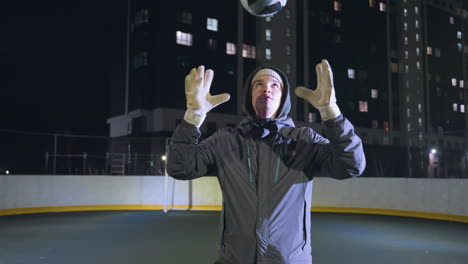 man in gloves throws a soccer ball into the air and catches it during an evening training session on an urban field, the illuminated background