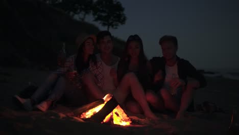 Young-cheerful-friends-sitting-by-the-fire-on-the-beach-in-the-evening,-singing-songs-and-drinking-beer-together.-Shot-in-4k