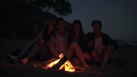 young cheerful friends sitting by the fire on the beach in the evening, singing songs and drinking beer together. shot in 4k