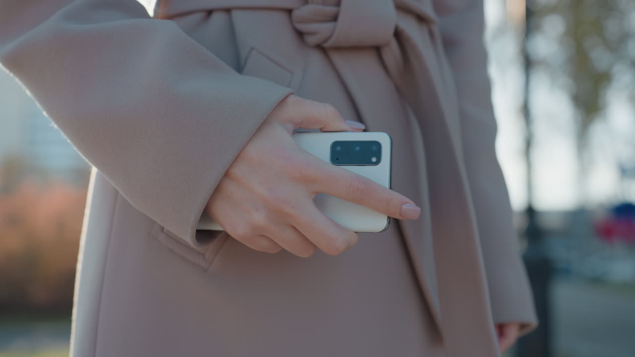Focused caucasian woman on phone, composed negotiation during city commute, beige coat and white scarf, professional manager closing deal, smartphone held at hip, sunlight rim light, pedestrian