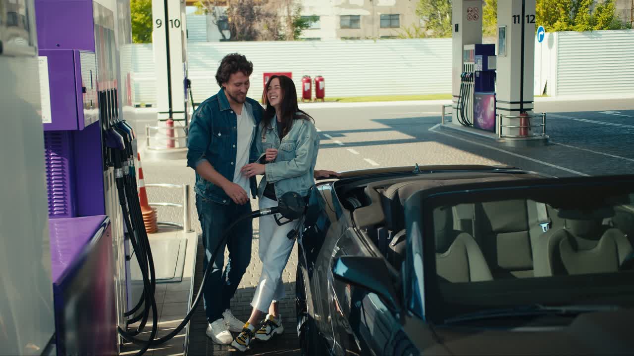 pareja feliz en la gasolinera: un chico y una chica en chaquetas de mezclilla reabastecen su descapotable gris oscuro, divirtiéndose y riendo