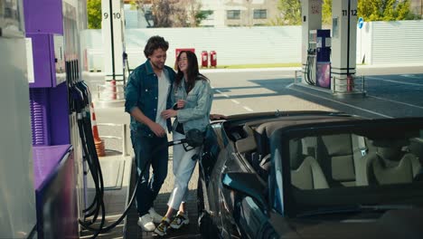 happy couple at the gas station: a guy and a girl in denim jackets refuel their dark gray convertible, having fun and laughing