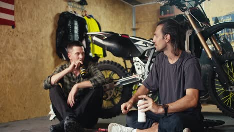 A male mechanic in a gray T-shirt makes gestures and tells his colleague about his vision of solving a problem during a rest and break at work. Repairing automobile equipment in a workshop