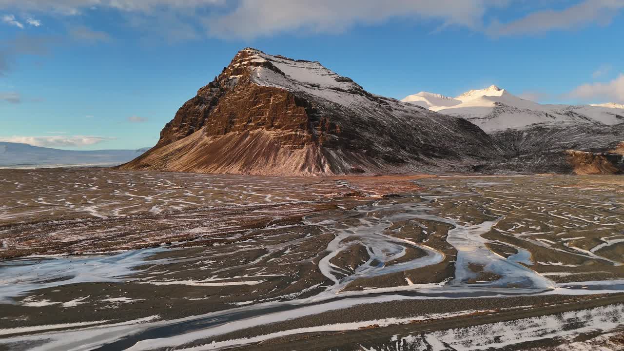Drone view of tall snow-capped mountain rising above frozen river plain with winding trails, scattered ice patches, and soft winter light under clear sky in remote wilderness of Iceland