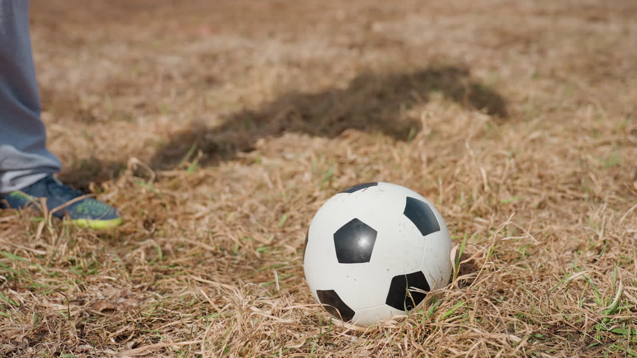 entorno de entrenamiento tranquilo con calzado cerca, escena iluminada por el sol que muestra equipamiento deportivo sobre una superficie de hierba seca, primer plano de un balón de fútbol apoyado junto a una zapatilla en hierba árida durante el entrenamiento
