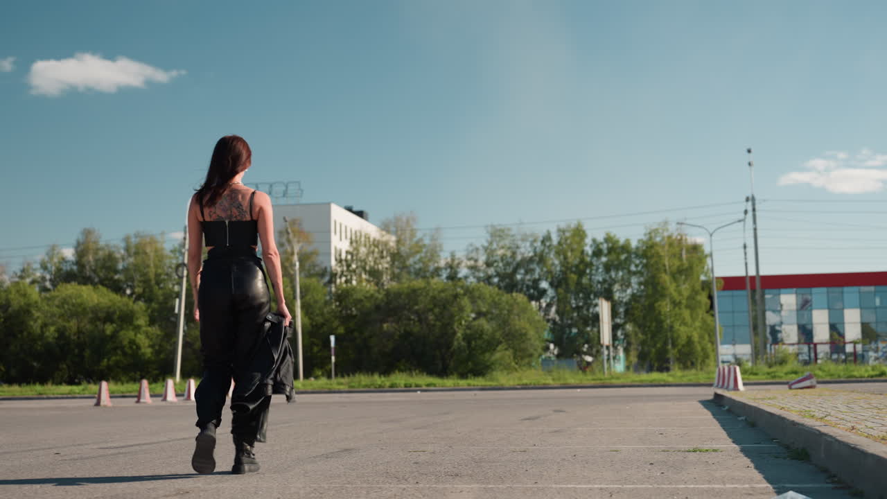 Rear view of female biker in black singlet walking confidently holding jacket in hand, tattoo visible on back, urban background with trees, buildings, and bright sky symbolizing strength