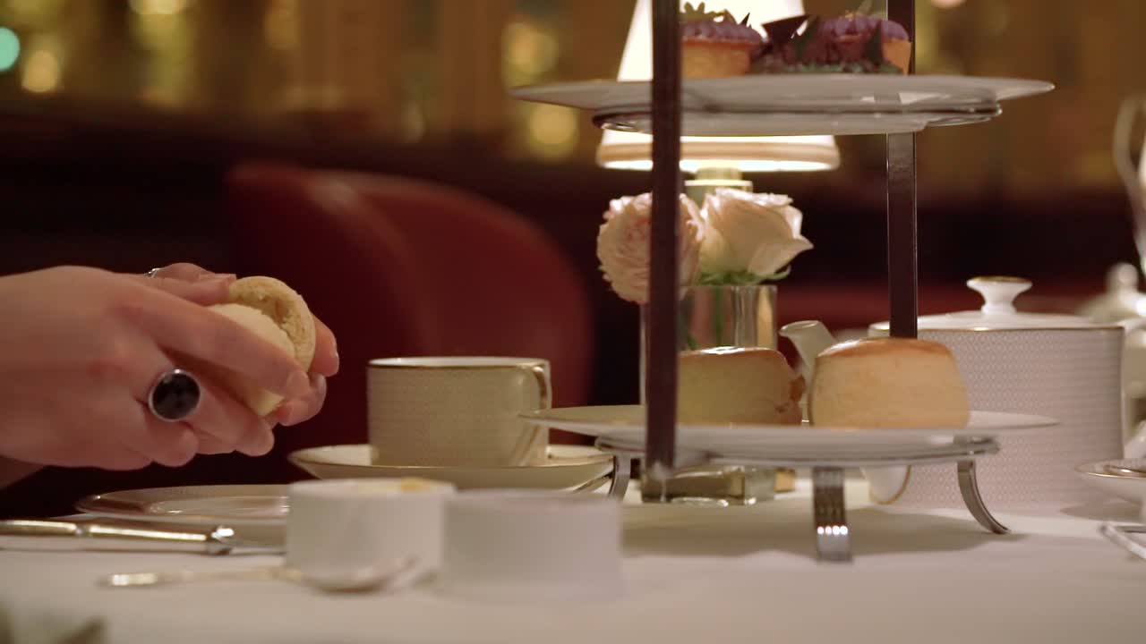 A lady's hand is picking up a piece of scone from the afternoon tea stand's lowest plate and tearing half. The camera is following her hand smoothly
