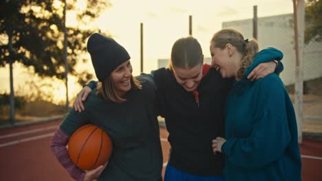 Close-up-a-trio-of-blonde-girls-in-sportswear-with-a-basketball-on-a-red-summer-court-on-the-street-stand-hugging-and-talking-after-their-match-in-the-morning-at-sunrise