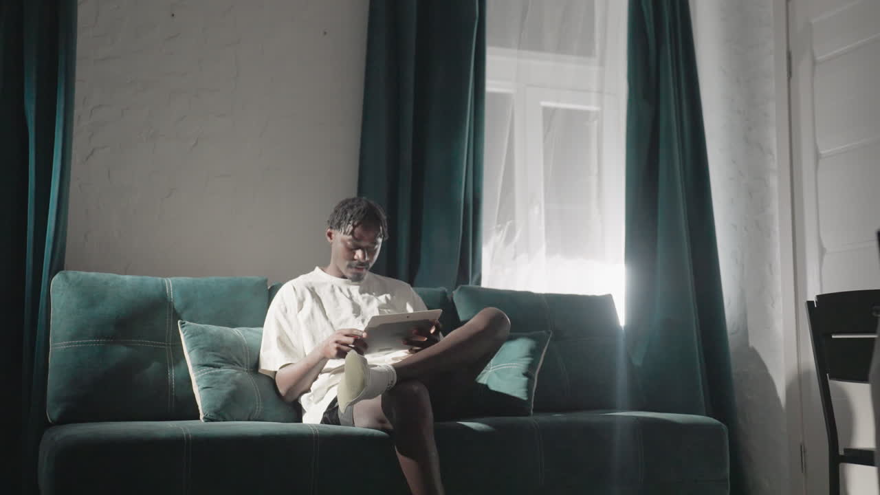 Champ sitting on sofa, reading tablet in cozy living room with teal curtains and soft lighting. Quiet, focused moment in serene home atmosphere, perfect for relaxation and mindfulness