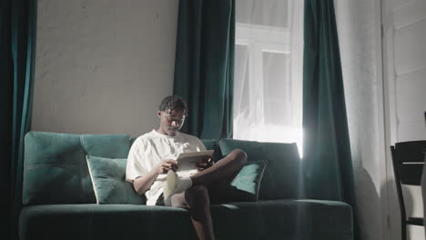 champ sitting on sofa, reading tablet in cozy living room with teal curtains and soft lighting. quiet, focused moment in serene home atmosphere, perfect for relaxation and mindfulness