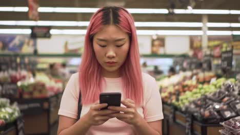 young woman with pink hair checking her phone in a vibrant grocery store aisle surrounded by fresh produce and vegetables, capturing everyday life moments