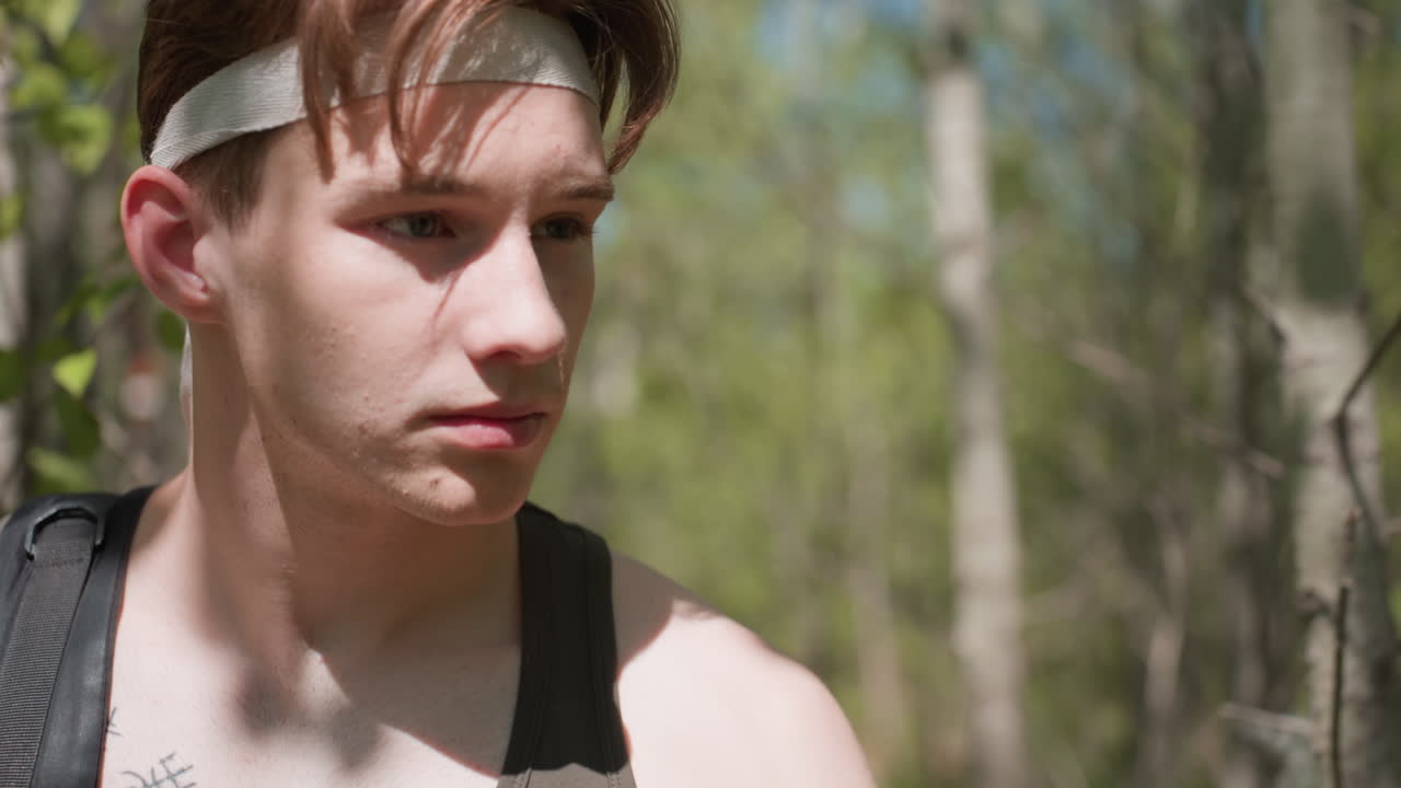 front view of tourist walking through dense forest undergrowth while dodging thin bare branch headband on forehead backpack strap on shoulder sunlight dappling fair skin and leaves swaying in breeze