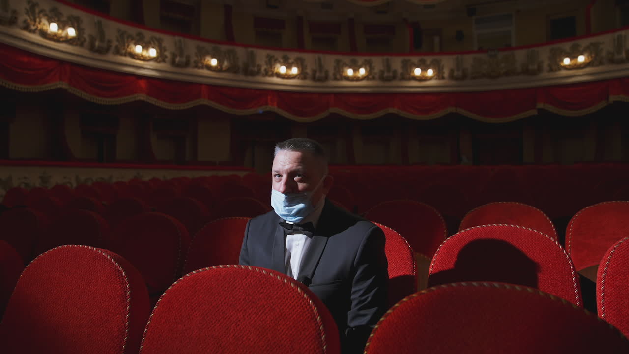 Alone businessman in mask in theater. Man in protective face mask sitting in a chair of empty auditorium. Theater in quarantine. Coronavirus pandemic.