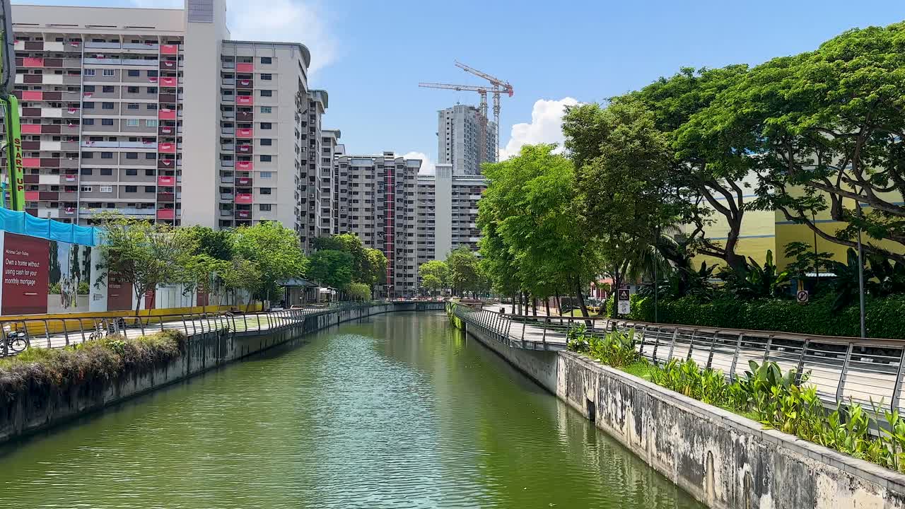 Slow camera pan along urban canal, high-rise apartments, greenery, bright daylight, clear blue sky