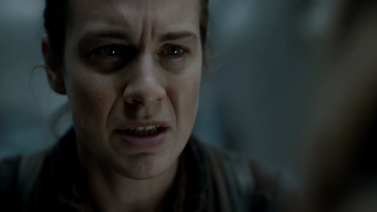Hearing offscreen voice, grimacing woman in dark jacket holding back tears in corridor, with straps