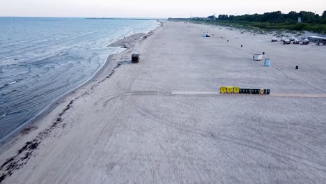 Luftaufnahme-Eines-Müllwagens,-Der-Sich-Am-Frühen-Sommermorgen-Nach-Sonnenaufgang-Am-Leeren-Strand-Entlang-Bewegt-Und-Sich-Auf-Die-Müllverladung-Vorbereitet,-Weitwinkel-Drohnenaufnahme,-Die-Sich-Vorwärts-Bewegt