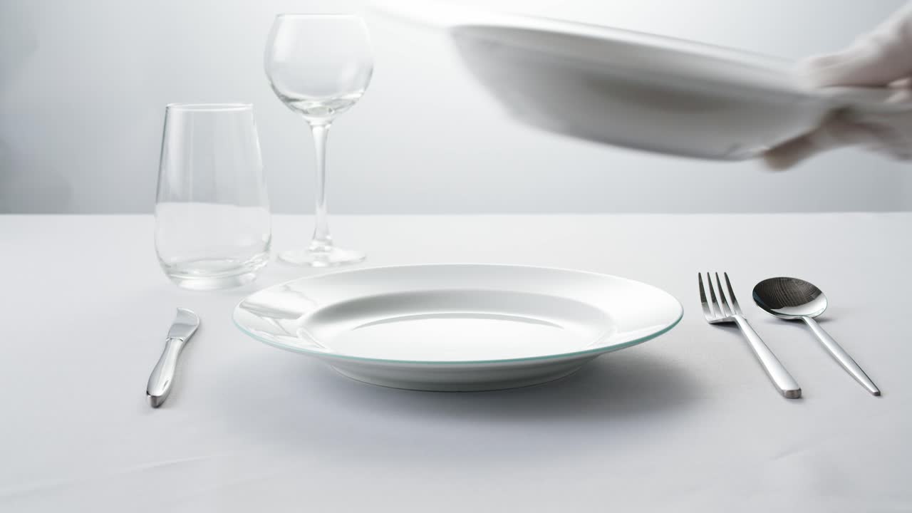 Professional table setting with white gloves. Plates, fork, knife, and glass arranged on a white table.