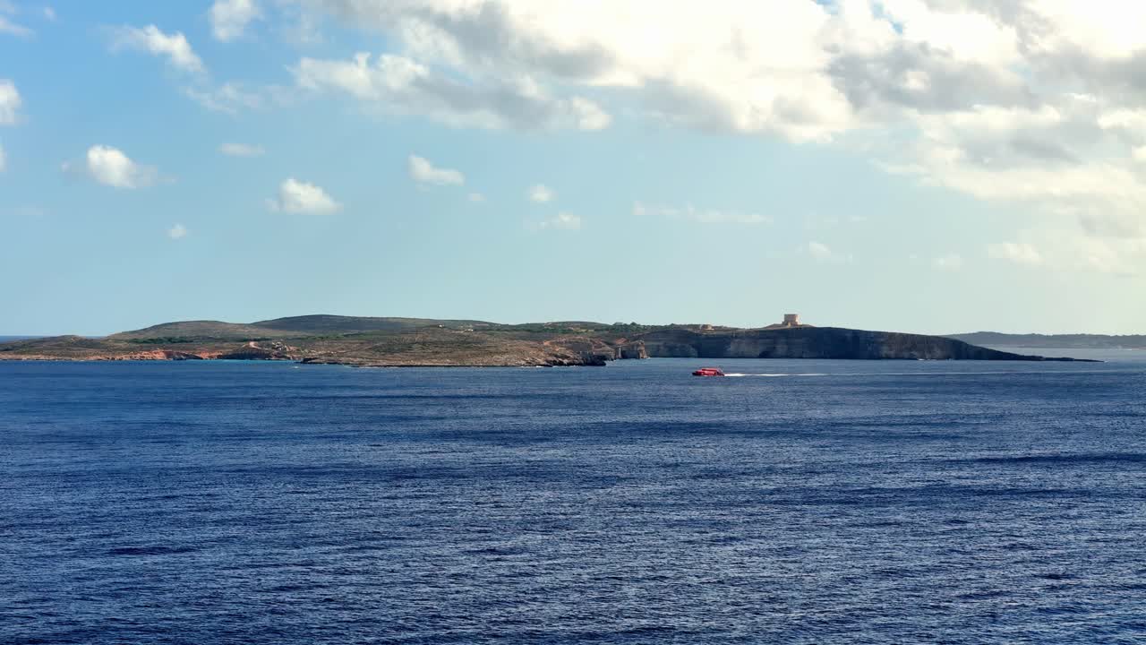 Rød passagerfærge, der sejler gennem den turkisblå North Comino Channel med det historiske Saint Marys Tower på de klippefyldte skrænter på Comino Island under en klar, solrig himmel på Malta om sommeren