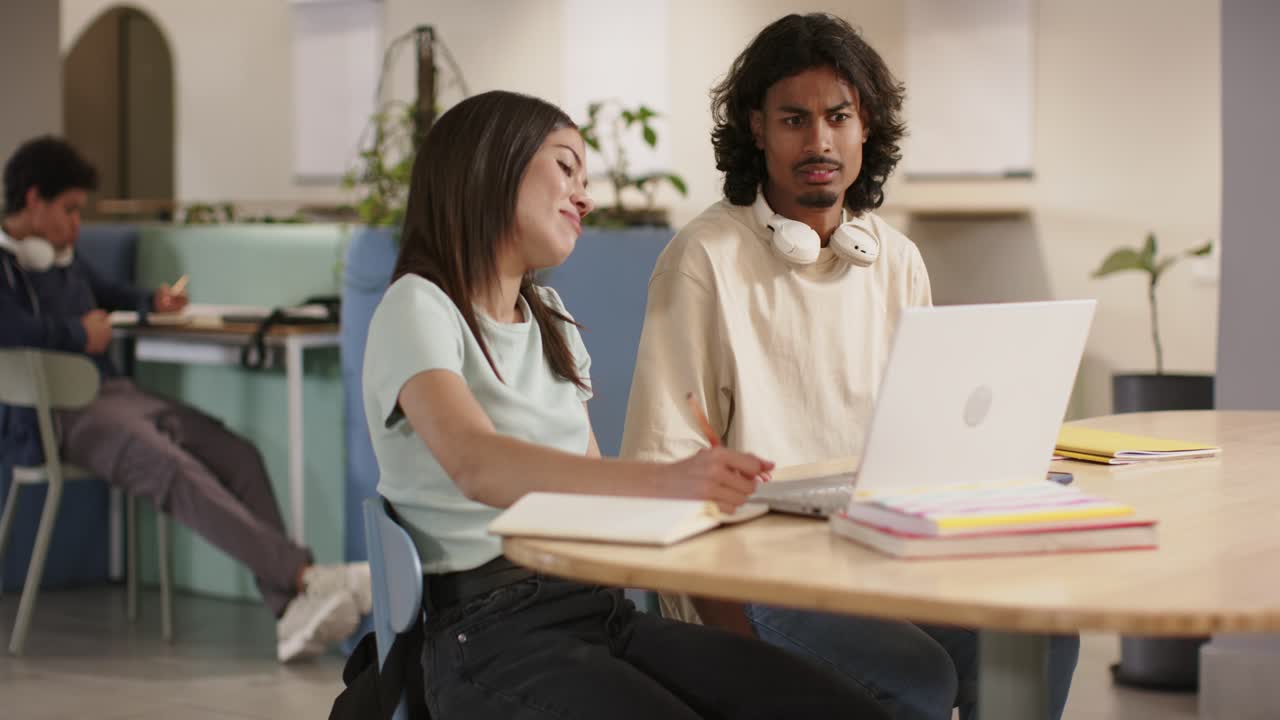 Two students working in study hub, man approaching to discuss laptop while woman writing notes