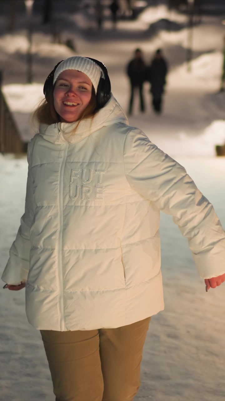 Woman wearing white puffer jacket and headphones moves through outdoor snowy environment possibly dancing or exercising. Her clothing includes word FUTURE on her coat and tan pants